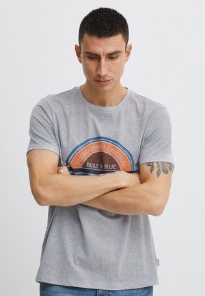 Moški s kratkimi temnimi lasmi in tetovažo na roki gleda navzdol, z rokami prekrižanimi, oblečen v sivo majico z napisom »Blend Denim Original Built in Blue«.