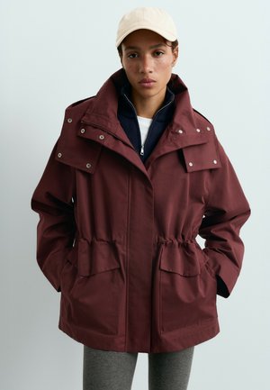 Femme portant une veste à capuche bordeaux avec des boutons-pression, une couche intérieure bleu marine zippée, un pantalon gris et une casquette de baseball beige, debout les mains dans les poches.