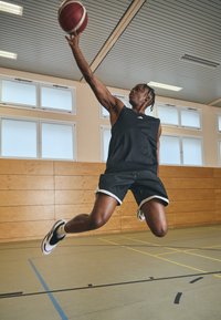 Basketbola spēlētājs melnā bezpiedurkņu krekla un šortus tērpā lecot, lai izmestu sarkano un baltu basketbola bumbu. Iekštelpu sports zāle ar koka sienām un pelēku grīdu.