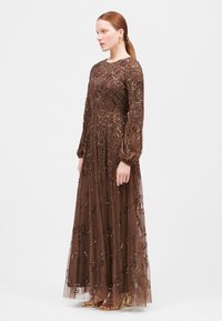 Bruine bloemengebroderde lange jurk met lange mouwen en transparante overlay, versierd met kralen en pailletten. Vloeiende silhouet, enkel-lang, gecombineerd met gouden sandalen.