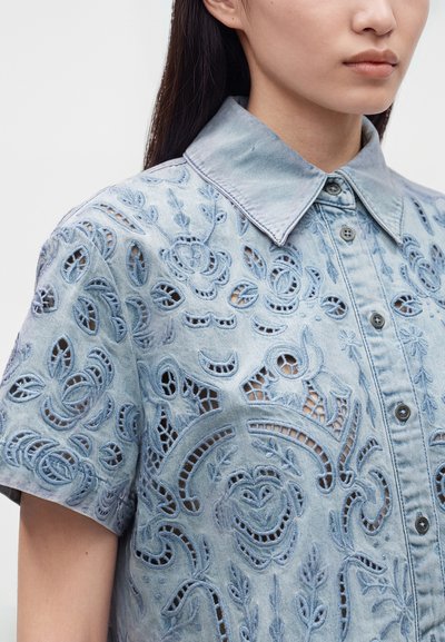 Femme portant une chemise en denim bleu clair à manches courtes avec une broderie florale ajourée complexe et un col pointu.