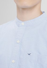 Light blue button-up shirt with a band collar, featuring a single pocket and a small embroidered logo. Made from a lightweight fabric.