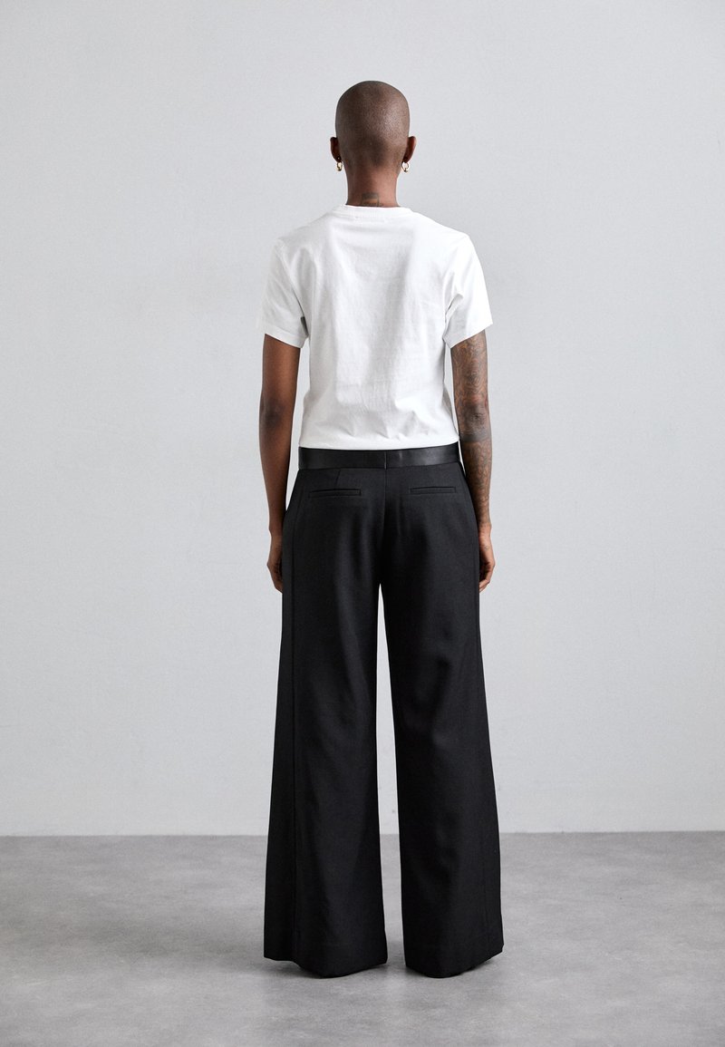 Camiseta de algodón blanca con mangas cortas, combinada con pantalones de pierna ancha negros que tienen bolsillos traseros y una textura suave, vista desde atrás.