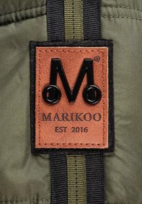Brown leather label with black embossed "M" and "MARIKOO EST 2016," sewn onto dark green fabric with black and olive striped accents.