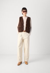 Brown faux shearling vest with two front pockets, featuring wooden toggles. Paired with light-colored, loose-fitting trousers and brown heeled boots.