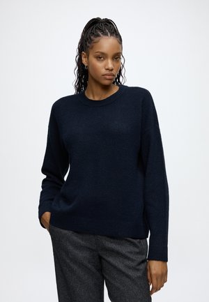 Femme aux cheveux tressés portant un pull bleu foncé et un pantalon gris, debout avec une main dans la poche sur un fond uni.