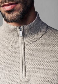 Lys grå strikket pullover med høj krave, halv-lynlås foran og struktureret mønster, der har en sølv lynlås trækdetaile.