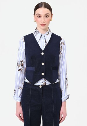 Donna con capelli scuri e riga centrale indossa orecchini d’oro, una camicia blu floreale a righe con bottoni, un gilet blu navy abbottonato e pantaloni abbinati.