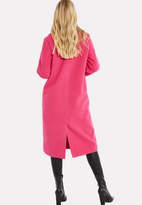 Donna bionda di spalle che indossa un lungo cappotto rosa acceso, leggings neri e stivali con tacco neri su uno sfondo bianco semplice.