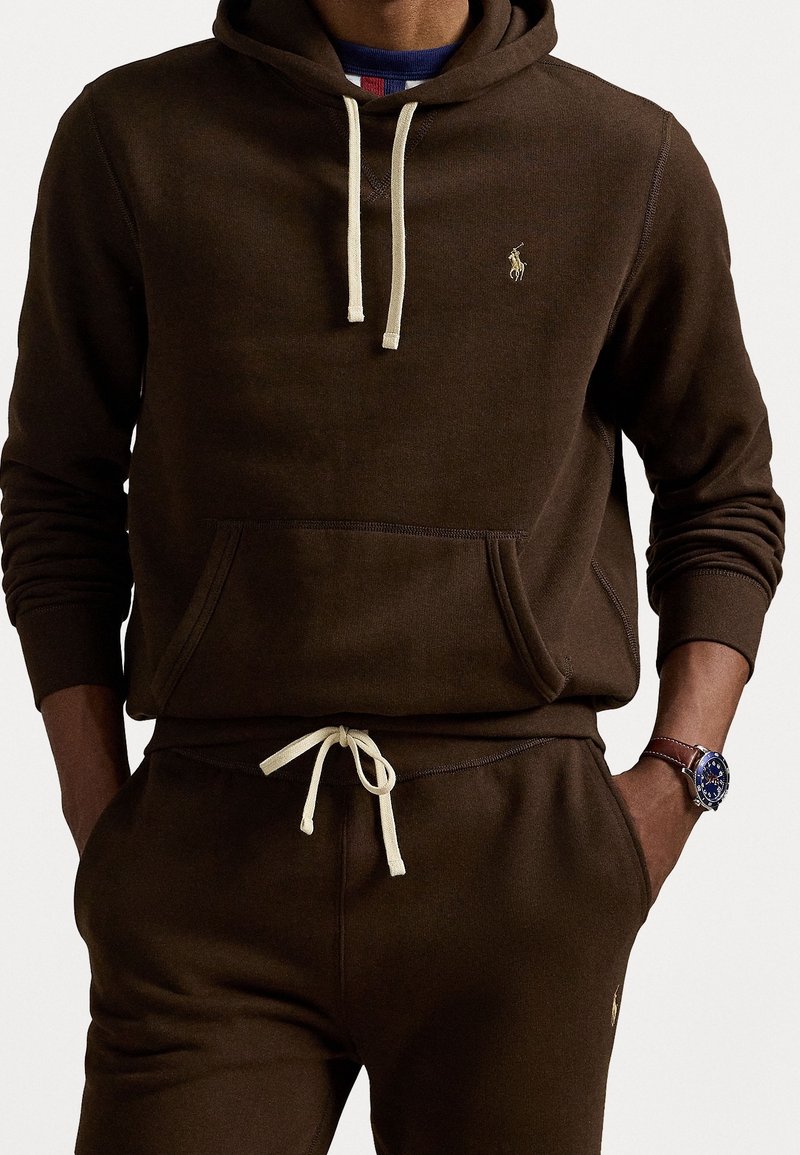 Homme portant un sweat à capuche marron foncé et un pantalon de survêtement assorti avec des cordons crème, ainsi qu'une montre avec un cadran bleu et un bracelet en cuir marron.