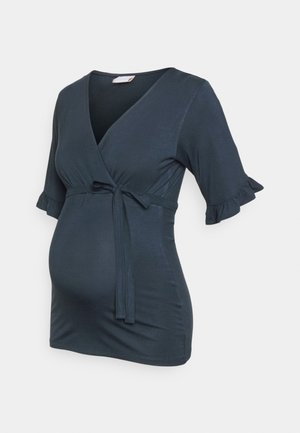 Donkerblauwe zwangerschaps top met V-hals, korte gerimpelde mouwen en een strik in de taille tegen een lichte achtergrond.