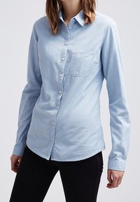 Ljusblå skjorta med knäppning, långärmad, framficka och en något böjd nederkant. Tyget ser mjukt ut med en texturerad yta.