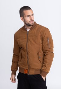 Bruine bomberjack met geribde manchetten en zoom, ritszakken en zichtbare merklolabel. Glad materiaal en gevoerd ontwerp voor warmte.