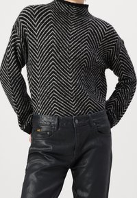 Suéter tejido en blanco y negro con patrón de zigzag y cuello alto, combinado con pantalones oscuros de apariencia cuero que presentan detalles texturales sutiles.