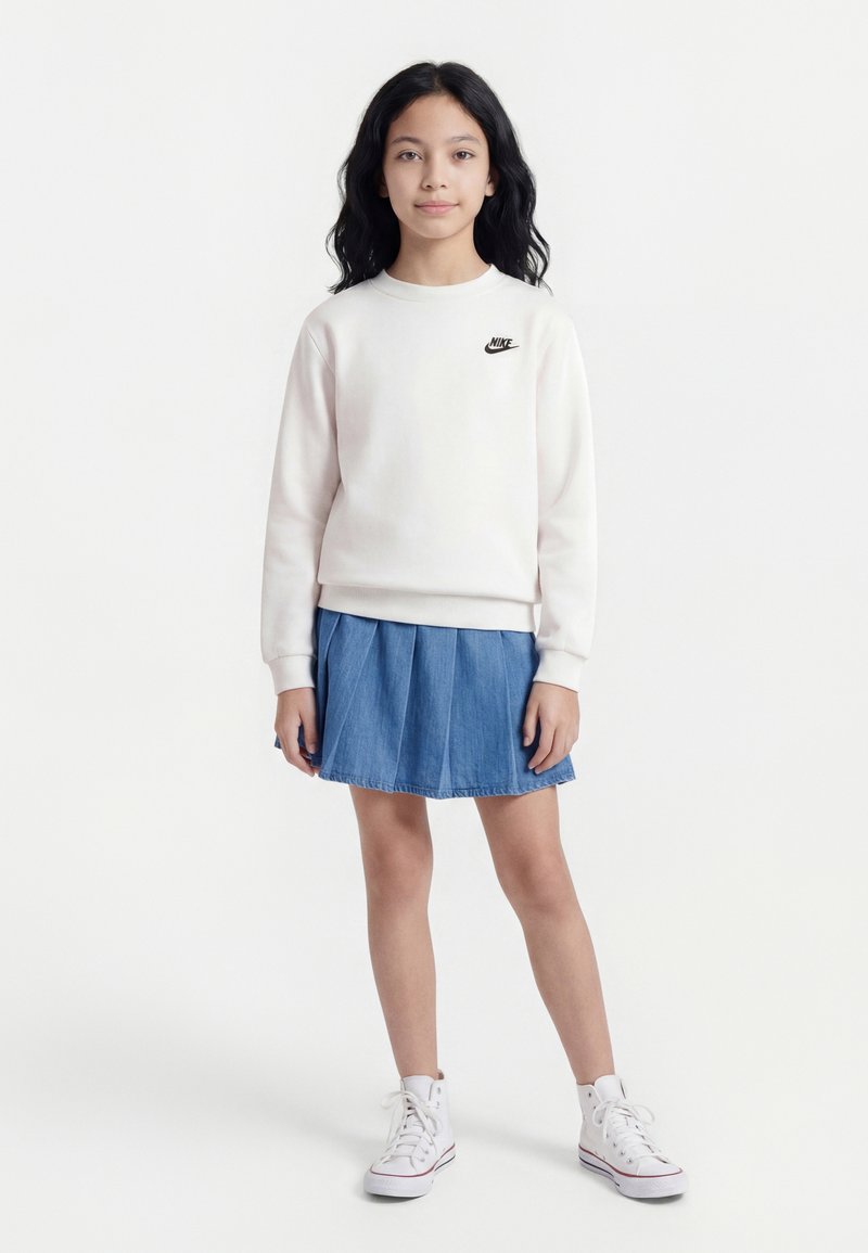 Νεαρή κοπέλα με μακριά σκούρα μαλλιά, φορεμένη με λευκό φούτερ Nike, μπλε πλισέ φούστα και λευκά αθλητικά παπούτσια ψηλής σόλας, στέκεται πρόσωπο προς τον φακό.