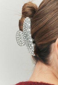 Silberner Haarklemmen mit Schmetterlingsform, verziert mit kleinen Strasssteinen, die braunes Haar in einem Dutt auf einem neutralen Hintergrund halten.