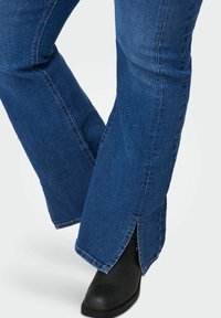 Jeans in denim blu a zampa d'elefante con uno spacco all'orlo, che mostra un boot nero sottostante. Il tessuto appare leggermente testurizzato.