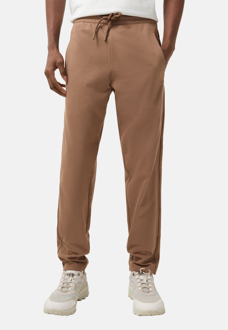 Brązowe dresy wykonane z miękkiej tkaniny, z pasem na sznurek, slim fit i bocznymi kieszeniami. Połączone z jasnymi butami sportowymi.