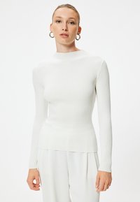 Haut blanc à manches longues et nervuré avec un col montant, fabriqué à partir d'un matériau doux. La coupe est légèrement ample au niveau de la taille.