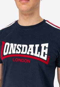 Námořnická modrá bavlněná trička s bílým a červeným textem "ONSDALE LONDON", s červenými a bílými pruhovanými akcenty na ramenech.