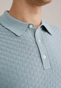 Svetlo modra pletena polo majica z teksturiranim šahovskim vzorcem, mehko ovratnico in dvema belima gumboma pri vratnem izrezu.