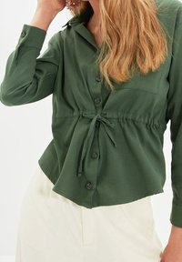 Vrouw gekleed in een olivegroen blouse met lange mouwen en knopen, met een tailleband en crèmekleurige broek, terwijl ze een bloemenhaarclip aanpast.