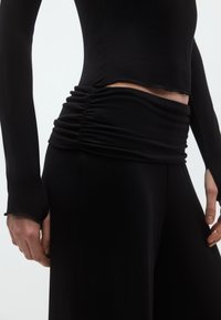 Μαύρο τοπ με μακριά μανίκια, με κοντό μήκος και φθαρμένα τελειώματα· ρυτιδωτή λεπτομέρεια στη μέση που δημιουργεί υφή και πτυχώσεις. Λείο ύφασμα.