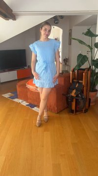 Γυναίκα με μπλε φόρεμα που στέκεται σταυροπόδι σε ένα σαλόνι με κόκκινο καναπέ, διπλωμένο όργανο κωπηλασίας, μεγάλο φυτό και τηλεόραση.