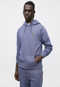 Lilla bomulds-hoodie med kængurulomme, justerbar snøre i hætten og gul broderet sommerfuglelogo på brystet, kombineret med matchende bukser.