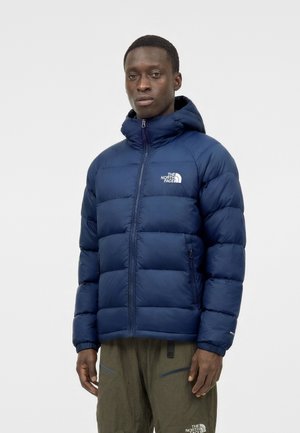 Modèle masculin portant une doudoune bleu marine avec capuche et fermeture éclair à l'avant, associée à un pantalon vert olive, debout devant un fond blanc uni.