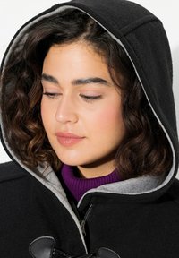 Cappotto di lana nera con cappuccio foderato grigio, dotato di zip e chiusure a toggle. Sotto è visibile un dolcevita a coste viola.