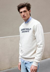 Creme sweatshirt med "SAINT-MALO UNIVERSITÉ" broderet i mørkeblåt. Båret over en lys blå stribet skjorte med krave og lys blå jeans.