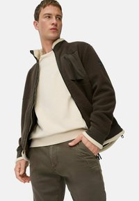 Brun fleecejakke med en cremefarvet foring, lynlås foran og en brystlomme. Båret over en beige sweatshirt og mørkegrønne bukser.