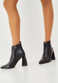 Botas de cuero negro hasta el tobillo con punta afilada, detalle de cremallera en el lateral y un tacón alto y grueso. Textura suave y diseño minimalista.