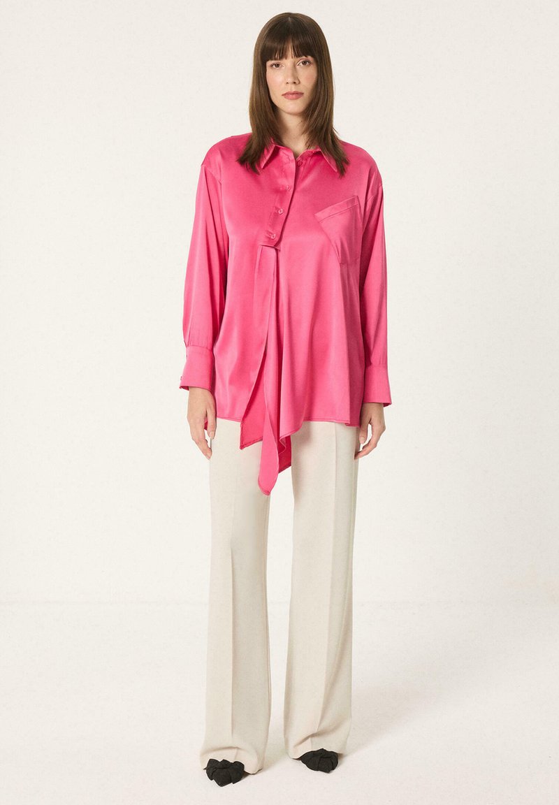 Femme debout portant un chemisier ample en satin rose avec un ourlet asymétrique et un pantalon large crème sur fond blanc.