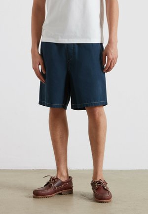 Mężczyzna ubrany w granatowe szorty, białą koszulkę i brązowe skórzane buty żeglarskie stoi na betonowej podłodze przy białej ścianie.