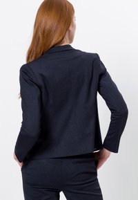Blazer bleu marine ajusté en tissu texturé avec col classique, dos ouvert et manches longues, assorti à un pantalon coordonné.