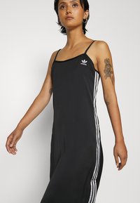 Črna športna obleka s tankimi naramnicami, okroglo vratno odprtino in belimi navpičnimi črtami ob straneh. Adidasov logotip na prsih.