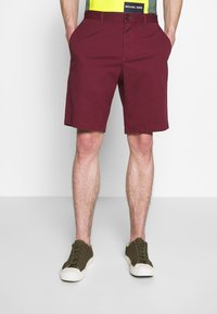 Pantaloni corti bordeaux in cotone, con tasche laterali, chiusura a bottone e orlo dritto, abbinati a sneaker verde scuro.