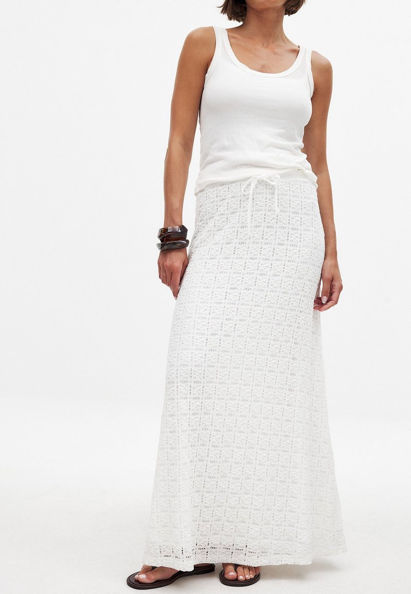 Jupe maxi blanche longue avec un motif en dentelle texturée et une taille à cordon. Portee avec un débardeur blanc et des sandales noires.