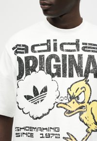 Κοντινό  πλάνο σε λευκό μπλουζάκι με φθαρμένο μαύρο κείμενο "adidas ORIGINAL" και ένα κίτρινο καρτούν πάπιο που κοιτάζει θυμωμένα ένα λογότυπο adidas μέσα σε μια φούσκα σκέψης.