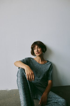 Graues, glitzerndes Outfit mit kurzen Ärmeln und lockerer Passform. Das Model sitzt vor einer neutralen Wand und präsentiert die Textur und den Glanz des Stoffs.