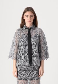 Spitzenbluse in Silber und Schwarz mit einem Kragen, halben Ärmeln und einem schwarzen Bindeband; florales Muster entlang der gesamten Bluse.