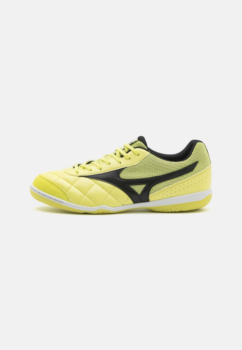 Zapatilla atlética de color amarillo lima con textura acolchada, acentos negros y parte superior de malla. Presenta una puntera redonda, cierre de cordones y suela de goma.