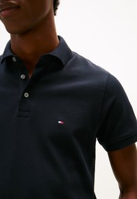 Vyras dėvi tamsiai mėlyną polo marškinėlį su mažu raudonos, baltos ir mėlynos spalvos vėliavos logotipu ant krūtinės, stovintis priešais vientisą foną.