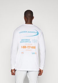 Valge pikkade varrukatega särk, mille seljal on sinise ja oranži tekstiga "Under Armour" logo koos kontaktandmete ja muude brändingute detailidega.
