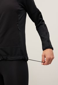 Giacca atletica nera in tessuto testurizzato, con tasche laterali con zip e lacci regolabili nell'orlo. Primo piano di una mano che regola il laccio.