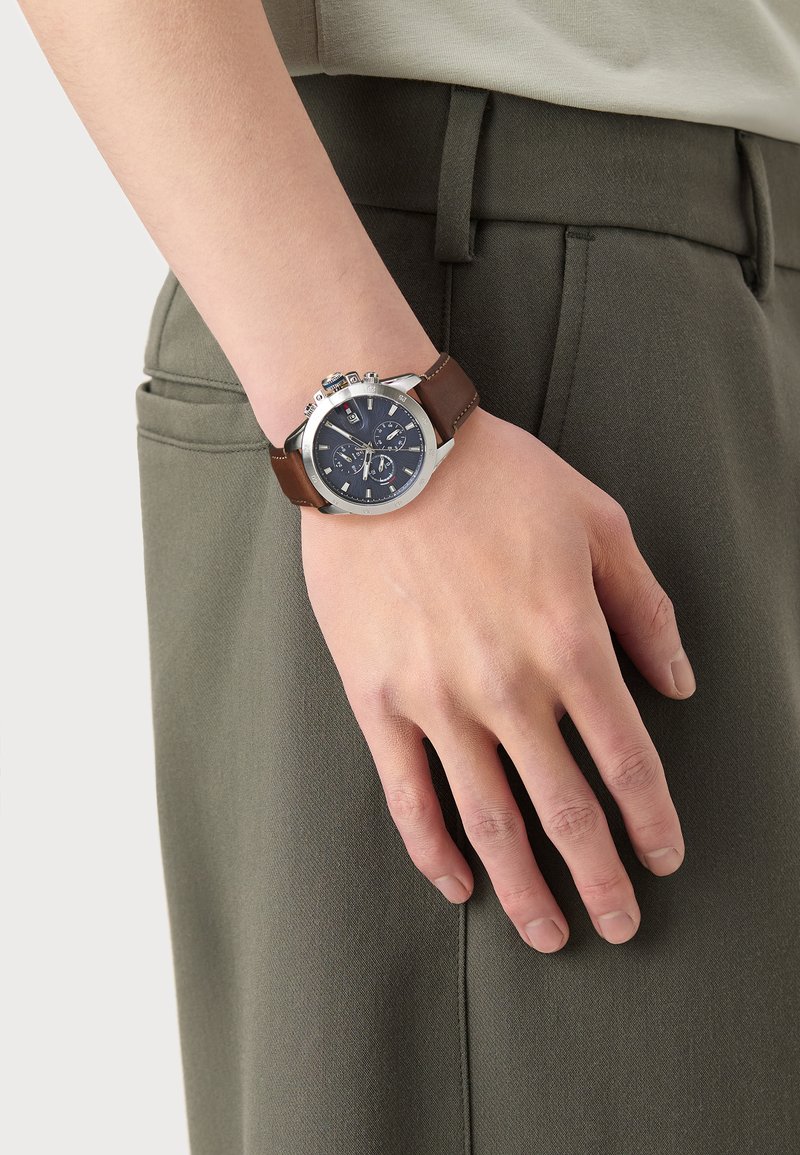 Montre en métal argenté avec un cadran bleu foncé, un bracelet en cuir marron, des fonctionnalités de chronographe et des accents argentés. Portée sur une main reposant sur un tissu vert.