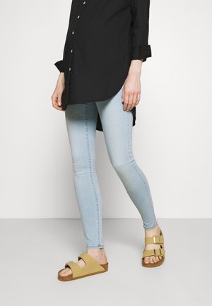 Personne portant un jean skinny bleu clair, un t-shirt noir à manches longues et des sandales beige à boucle, debout devant un fond uni.