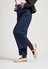 Pantalones de mezclilla oscuros de cintura alta, con piernas anchas y costuras blancas, combinados con zapatos planos beige y calcetines burdeos para un contraste.